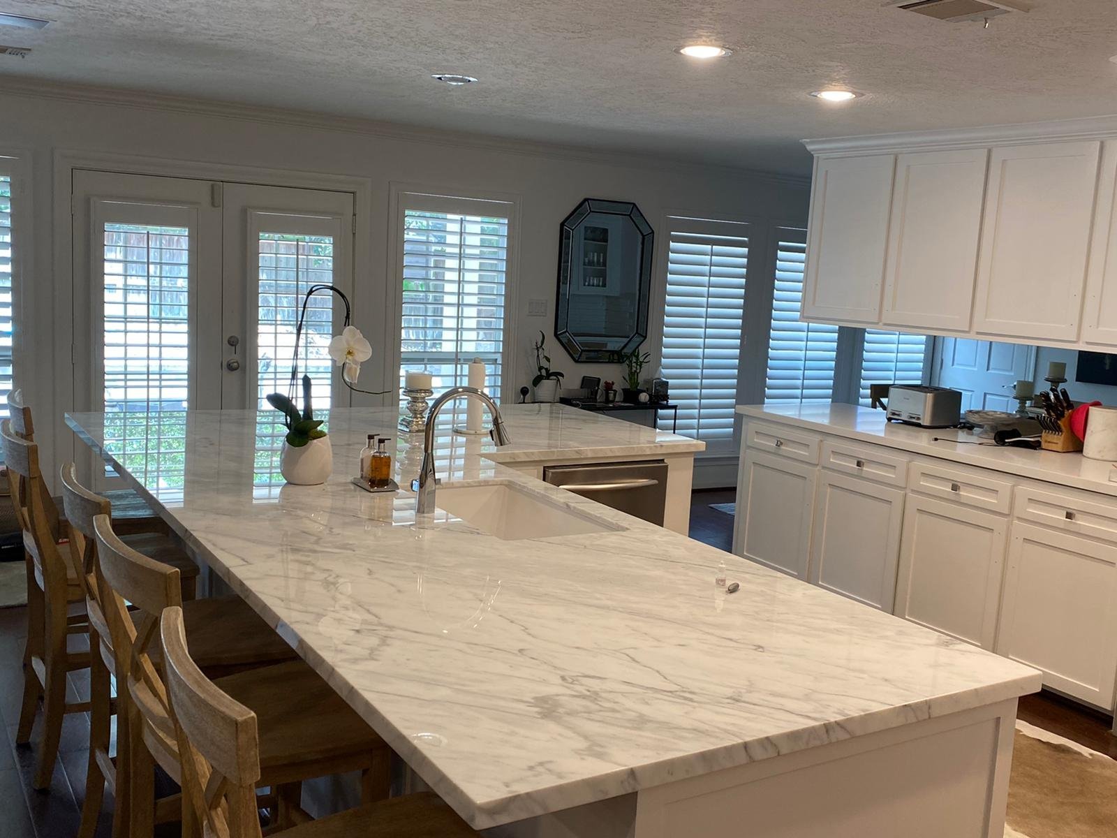 Modern kitchen with marble waterfall island, white cabinetry, and floor-to-ceiling shuttered windows overlooking water.