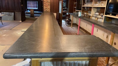 Long dark concrete countertop in modern kitchen with exposed brick and shelving in background