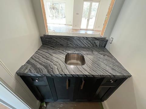 Black bathroom vanity with gray marble countertop and stainless steel sink, viewed from above in a bathroom hallway
