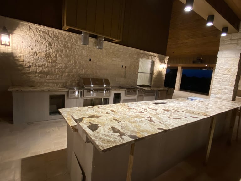 Modern luxury outdoor kitchen at night with granite countertops, stainless steel grills, and white painted brick walls illuminated by overhead lighting